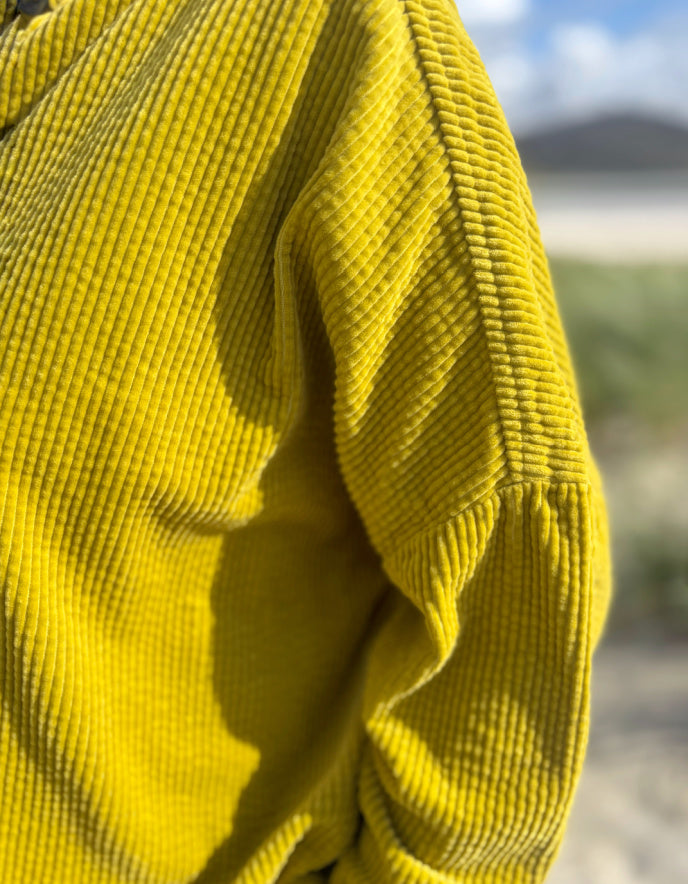 Close-up of a bright yellow ribbed knit garment, likely a sweater or jacket sleeve, with a textured corduroy-like rib pattern and a softly blurred outdoor background.