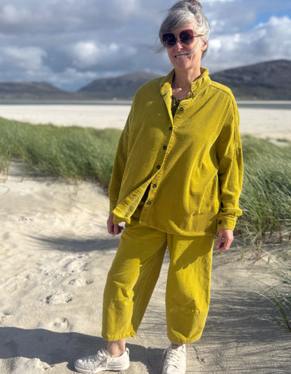 A woman wearing a matching mustard yellow corduroy button-up shirt and pants set stands on a sandy beach with dune grass and mountains in the background.