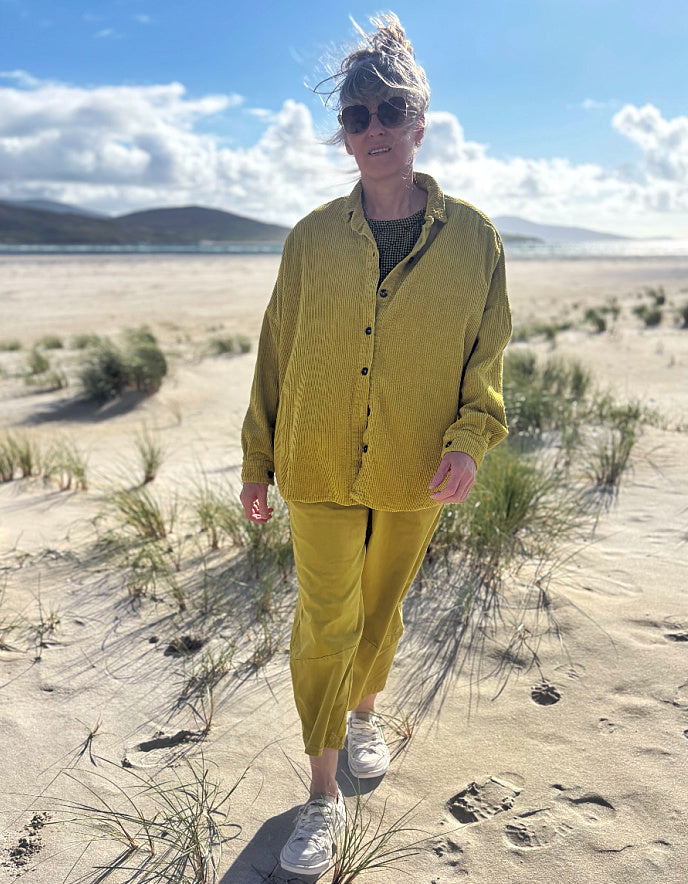 A woman walking on a sandy beach wearing a matching olive yellow corduroy button-up shirt and trousers set, white sneakers, and sunglasses.