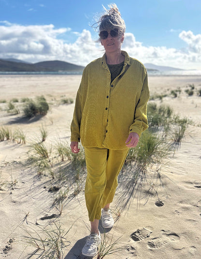 A woman walking on a sandy beach wearing a matching olive yellow corduroy button-up shirt and trousers set, white sneakers, and sunglasses.