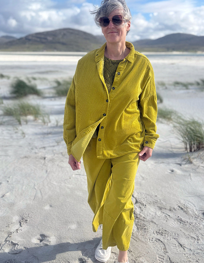 A woman with short gray hair and sunglasses walking on a sandy beach wearing a matching yellow corduroy button-up shirt and pants set over a green patterned top.