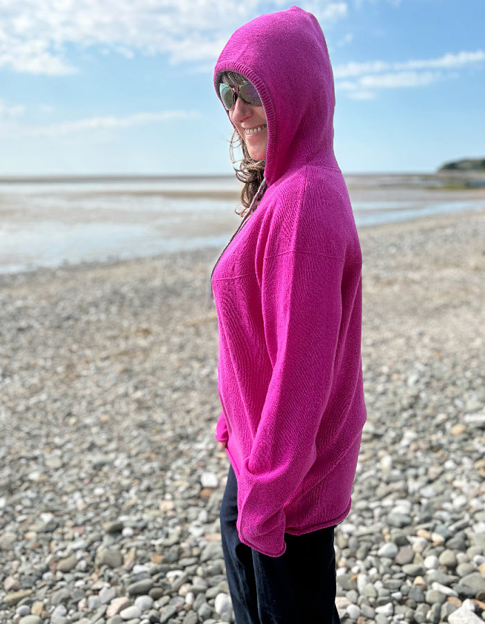 Person wearing a bright pink hoodie on a pebbly beach with a blue sky.