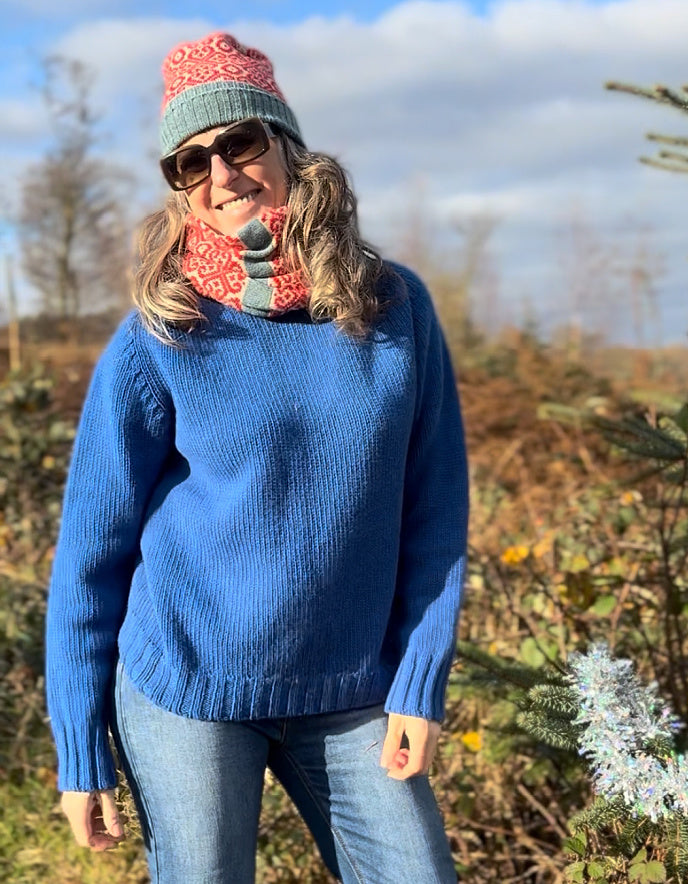 Person wearing a blue sweater, colorful hat, and scarf outdoors with trees and sky in the background