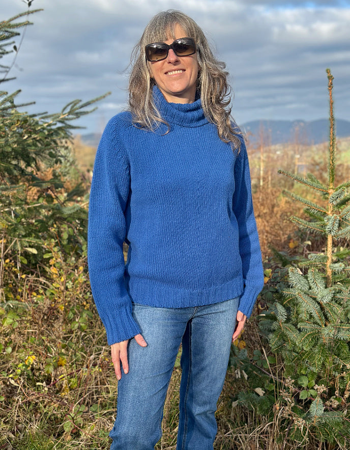 Person wearing a blue sweater, colourful hat, and scarf outdoors with trees and sky in the background