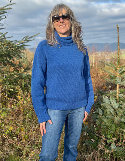 Person wearing a blue sweater, colourful hat, and scarf outdoors with trees and sky in the background