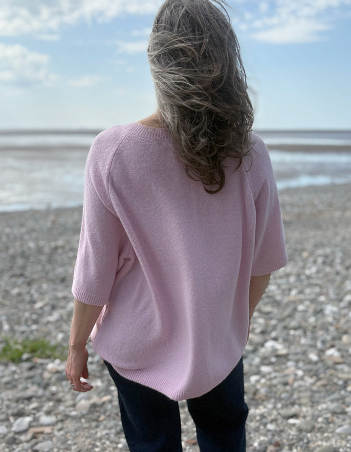 Person wearing a pink sweater standing on a pebbly beach with a blue sky.