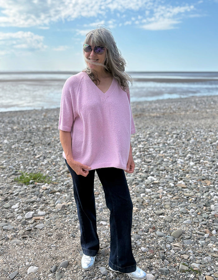 Woman in a pink sweater and black pants standing on a pebbly beach with blue sky and ocean in the background.