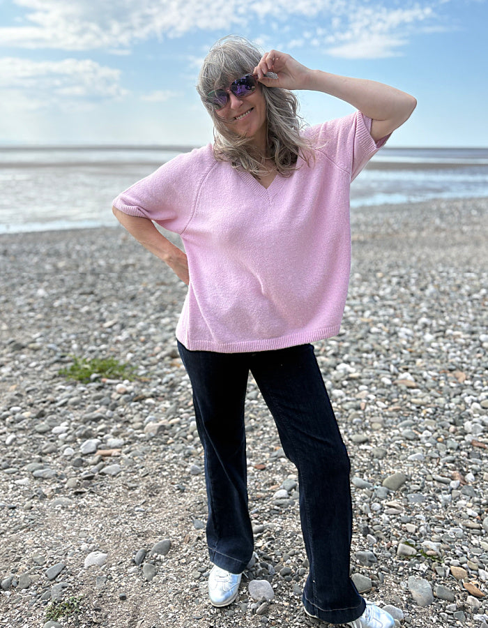 Person wearing a pink sweater and black pants standing on a pebbly beach.