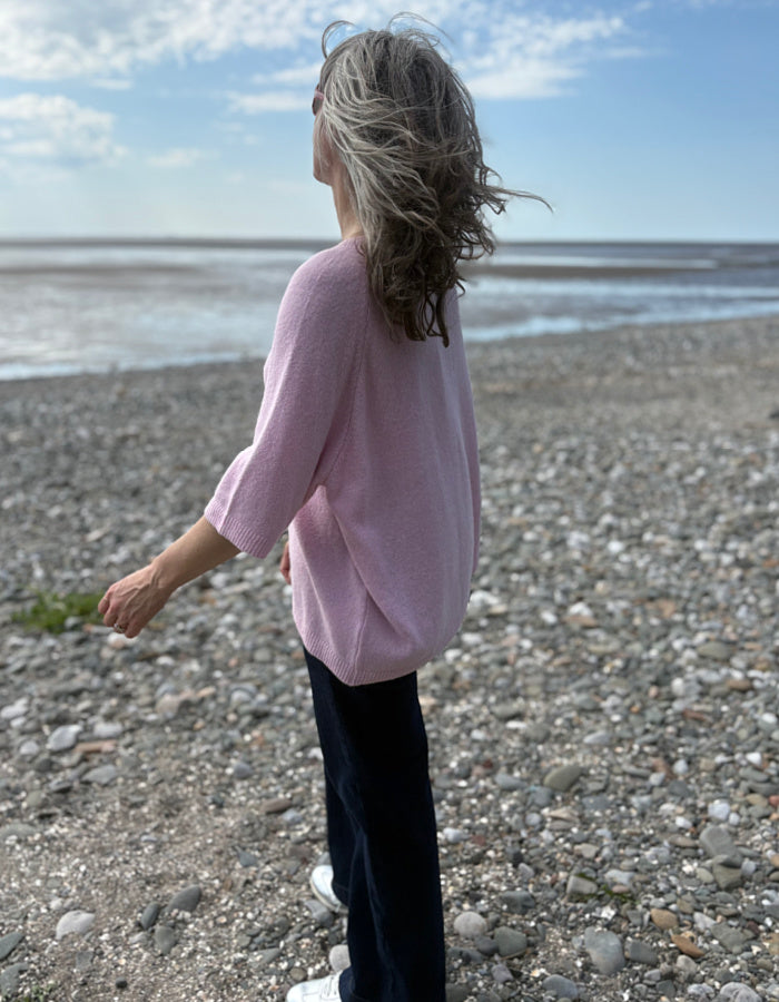 Person wearing a pink sweater standing on a pebbly beach with a blue sky.