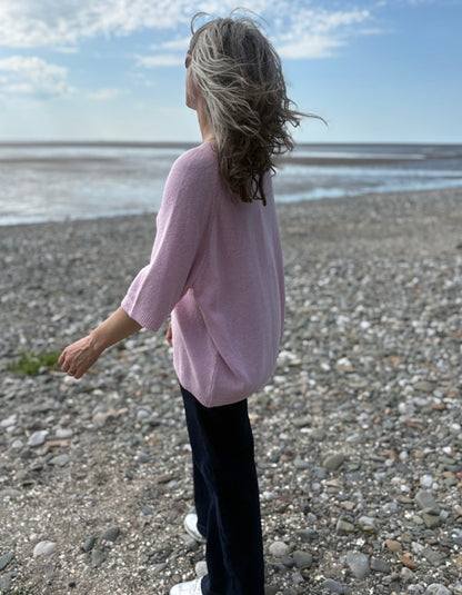 Person wearing a pink sweater standing on a pebbly beach with a blue sky.