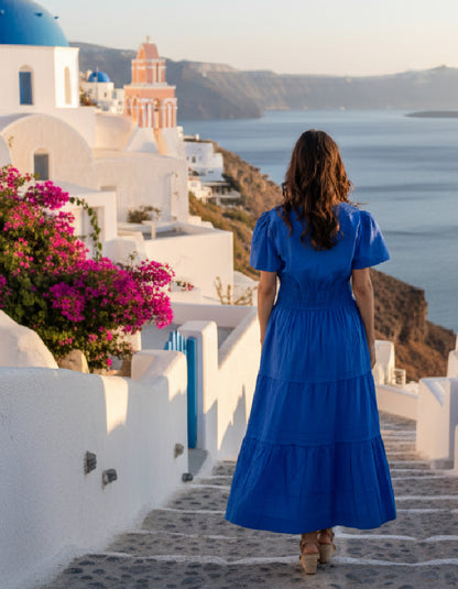 Woman in a blue dress standing on a scenic pathway with white buildings and ocean view.