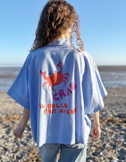 Person wearing a light blue wide fit cotton shirt with a red crab design and text on a beach.