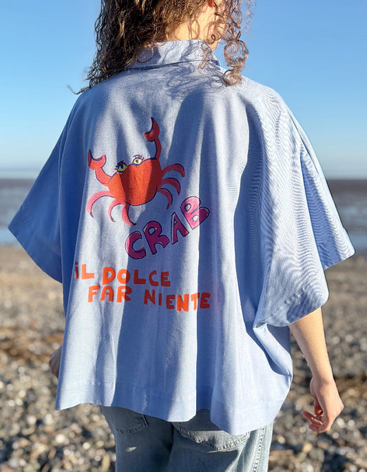 Person wearing a light blue wide fit cotton shirt with a red crab design and text on a beach.