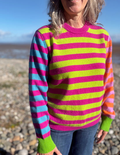 A woman wearing a vibrant knit sweater with bold horizontal stripes in neon pink, lime green, light blue, and orange, standing outdoors on a rocky beach.