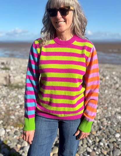 A woman wearing a vibrant, multicolored striped knit sweater with pink, lime green, orange, and blue horizontal stripes, standing on a rocky beach.