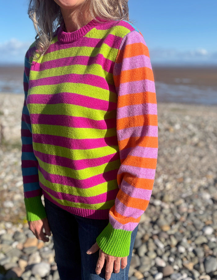 A person wearing a bright striped knit sweater with horizontal bands of pink, lime green, orange, and lavender, standing outdoors on a pebble beach.