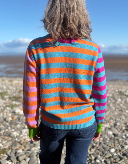 A person standing on a pebble beach, viewed from behind, wearing a fuzzy knit sweater with bold horizontal stripes in orange, turquoise, pink, purple, and green.