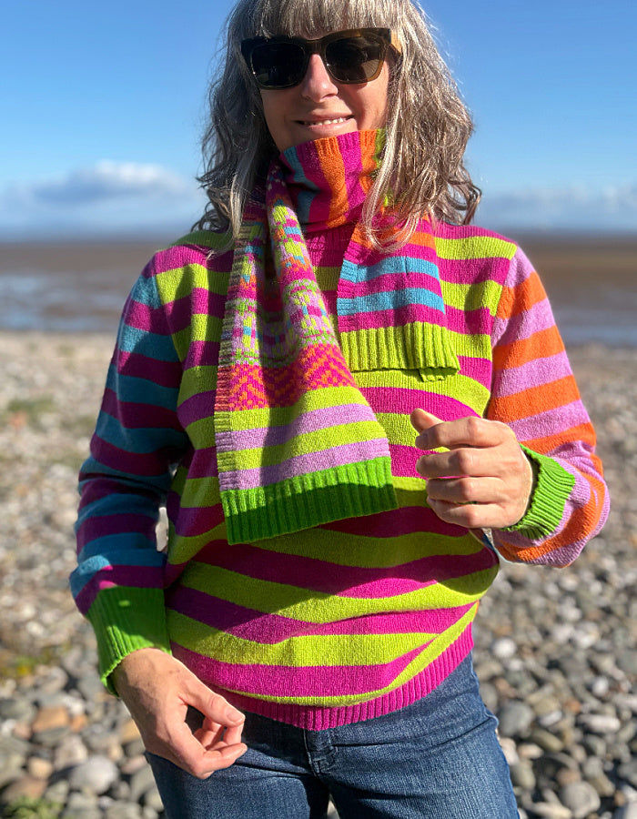 A woman wearing a vibrant, multicolored striped knit sweater and a matching striped knit scarf, standing outdoors on a rocky beach with water and sky in the background.