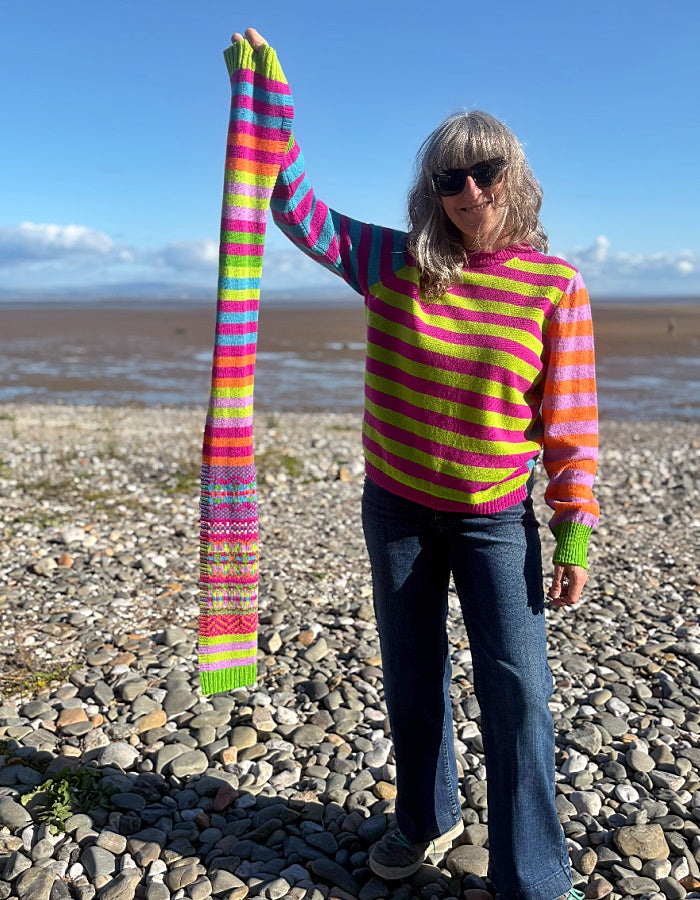 A woman standing on a pebble beach wearing a bright striped knit sweater and holding up a matching striped knit scarf or wrap. The garment has bold horizontal bands in pink, lime green, blue, orange, and yellow, with a patterned section near the lower end.