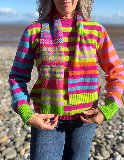 A woman wearing a vibrant, multicolored striped knit sweater and a matching striped knit scarf, standing outdoors on a pebble beach.