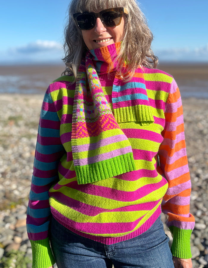 A woman wearing a vibrant, multicolored striped knit turtleneck sweater and matching knit scarf, standing outdoors on a pebble beach with water and sky in the background.