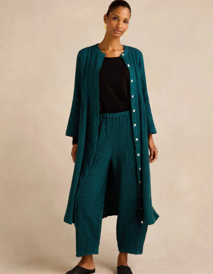 A woman modeling a matching teal green two-piece set consisting of a long button-front cardigan and wide-leg cropped pants, styled over a black top, against a plain beige studio background.