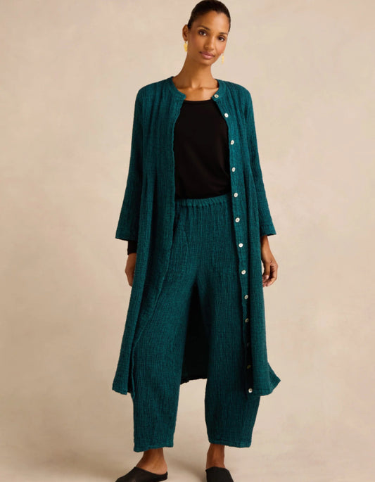 A woman modeling a matching teal green two-piece set consisting of a long button-front cardigan and wide-leg cropped pants, styled over a black top, against a plain beige studio background.
