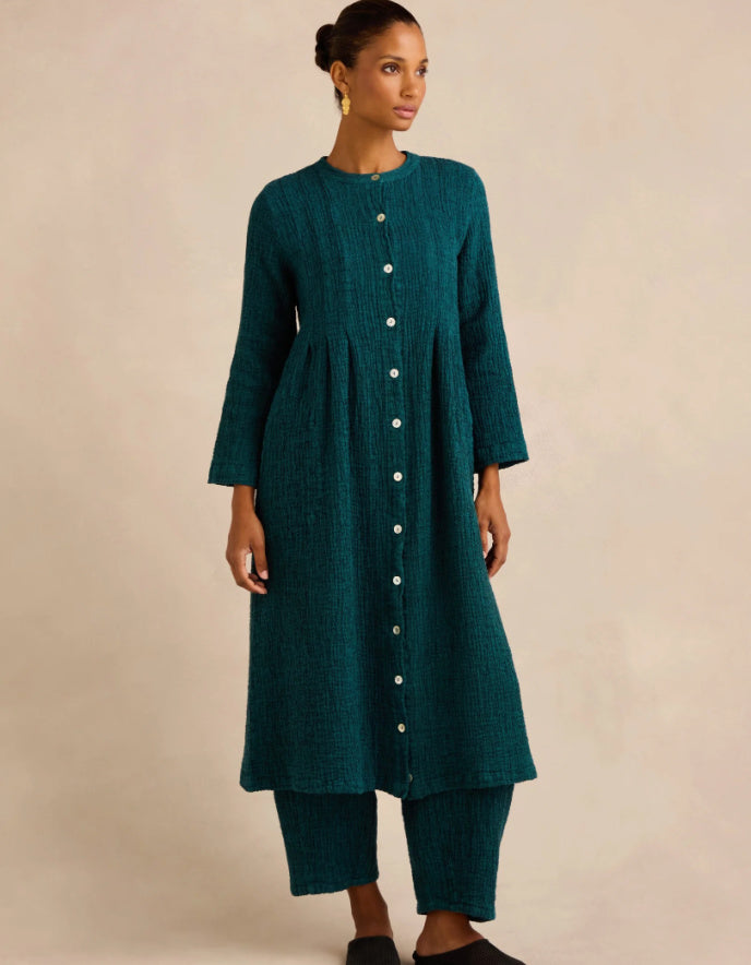 A woman modeling a dark teal green long-sleeved tunic with a button-down front, paired with matching cropped pants, against a plain beige studio background.
