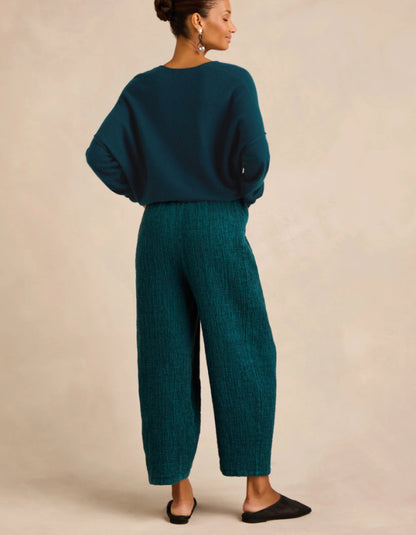 A woman shown from the back wearing a dark teal knit sweater and matching textured wide-leg trousers, paired with black flats, against a warm beige studio background.
