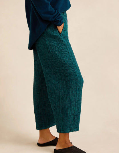 A person wearing textured wide-leg cropped pants in deep teal, paired with a navy long-sleeve top and black slip-on shoes, against a plain beige studio background.
