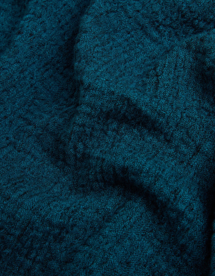 A close-up of a dark teal textured fabric, showing a detailed knit or woven pattern with soft folds and shadows.