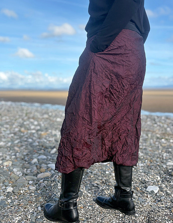 Person wearing a burgundy skirt and black boots standing on a pebbly beach with a blue sky.