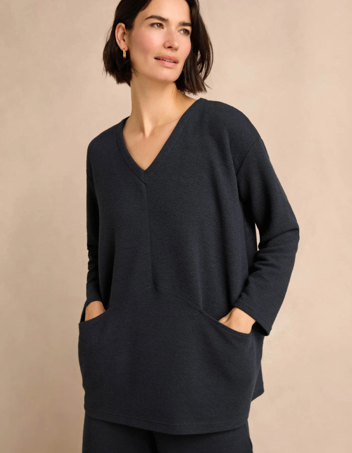 A woman wearing a navy V-neck long-sleeve tunic with side pockets, posed against a solid beige background.