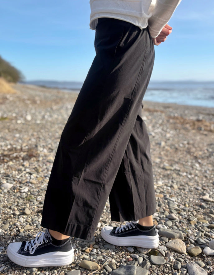 Person wearing black wide-leg pants and black sneakers on a pebbly beach.