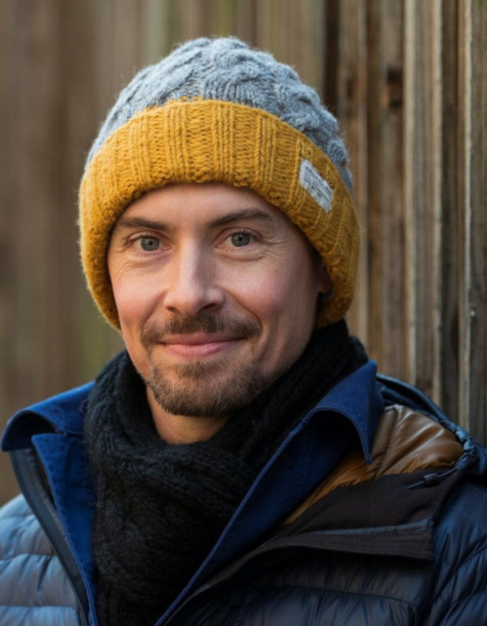 Man wearing a gray and yellow knit beanie with a blue jacket against a wooden background