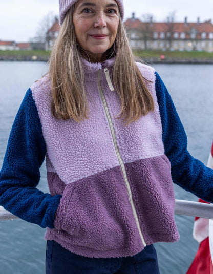 A woman wearing a color-blocked fleece vest with a lavender top section, a light pink middle band, and a dusty purple bottom section, paired with a matching lavender beanie, photographed outdoors by the water.