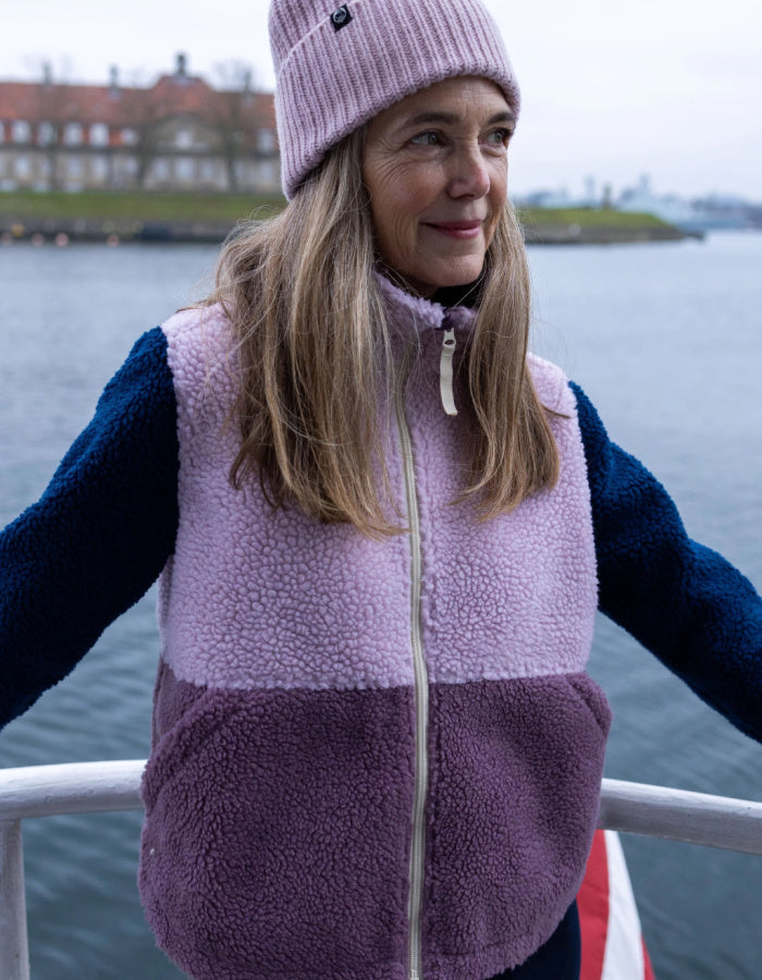 A woman wearing a two-tone fleece zip-up vest with a light purple upper section and a darker mauve-purple lower section, paired with a matching knit beanie, photographed outdoors by the water.