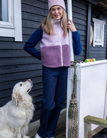 A woman wearing a colour-block fleece jacket in pink, purple, and navy blue stands on a wooden deck next to a white railing, with a white dog sitting beside her. The vest has a light pink top section, a darker purple middle section, and navy blue sleeves 