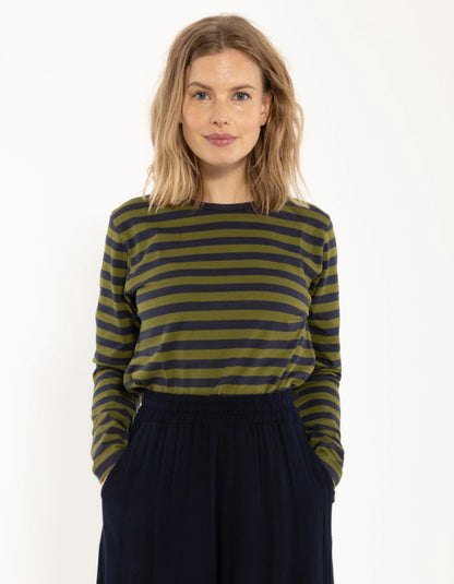 A woman with blonde hair wearing a long-sleeve top with olive green and black horizontal stripes, paired with dark navy wide-leg pants, standing against a plain white background.