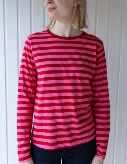 A woman wearing a long-sleeve crewneck shirt with horizontal pink and red stripes, paired with dark blue jeans, standing against a white paneled wall.