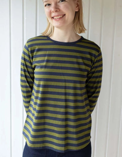 A woman wearing a long-sleeve crewneck shirt with horizontal olive green and navy blue stripes, standing against a white paneled wall.