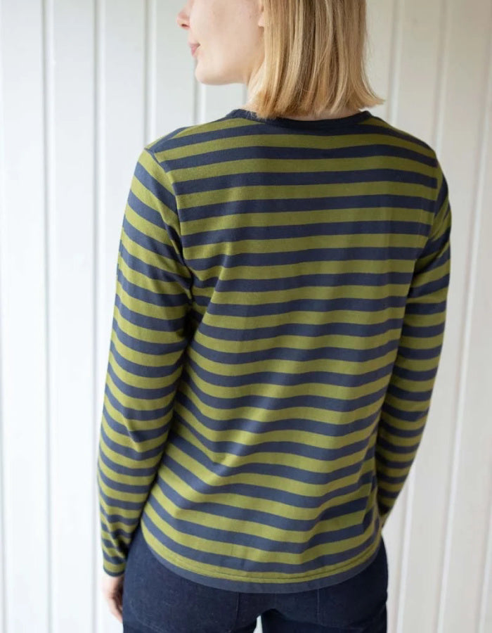 A woman shown from the back wearing a long-sleeve striped shirt with olive green and dark navy horizontal stripes, paired with dark blue jeans, set against a white paneled wall.