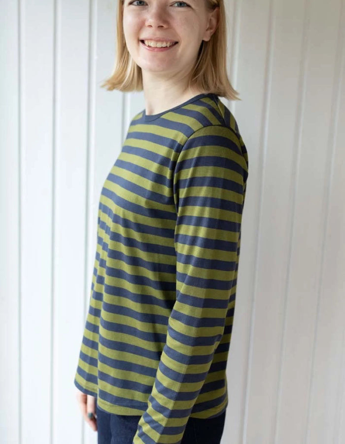 A woman wearing a long-sleeve crewneck top with horizontal olive green and navy blue stripes, standing against a white paneled wall.