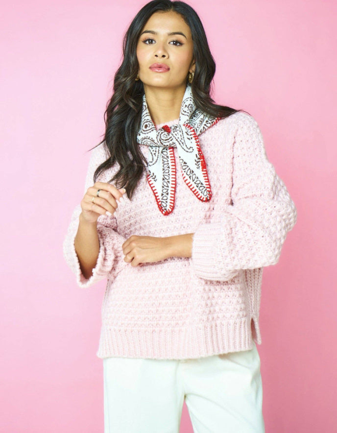A woman modeling a light pink textured knit sweater with a black, white, and red paisley bandana tied around the collar, set against a solid pink background.