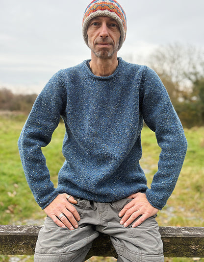 Man wearing a blue knitted sweater and colorful beanie standing outdoors with a natural background.