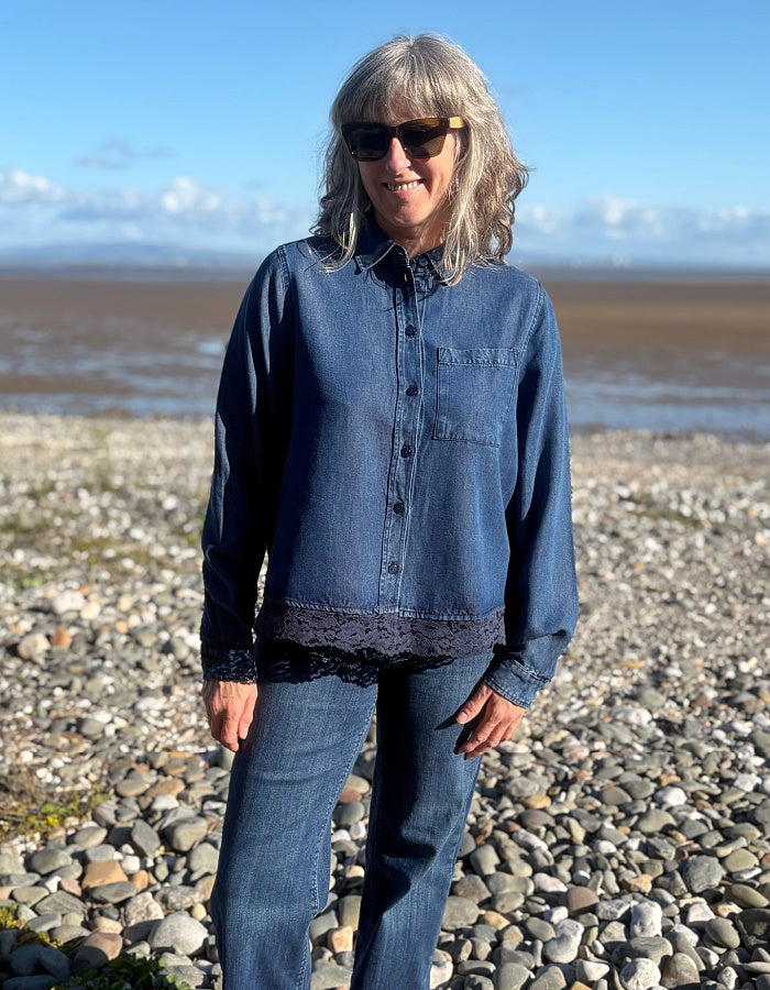A woman with gray hair and sunglasses stands on a pebble beach wearing a dark blue denim button-up shirt with a black lace hem detail and matching blue jeans.
