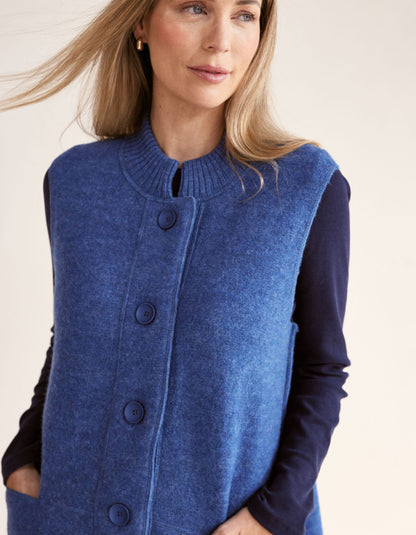Woman wearing a blue boiled wool long vest over a dark long-sleeve shirt on a light pink background