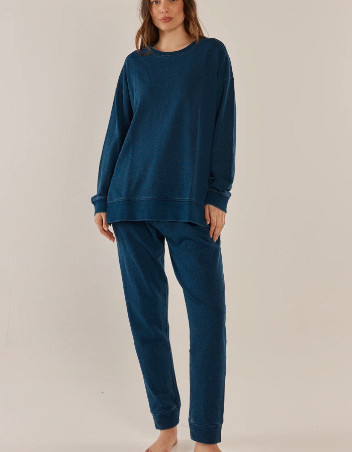 A woman modeling a matching dark blue lounge set consisting of a long-sleeve sweatshirt and jogger pants, photographed against a plain light beige studio background.
