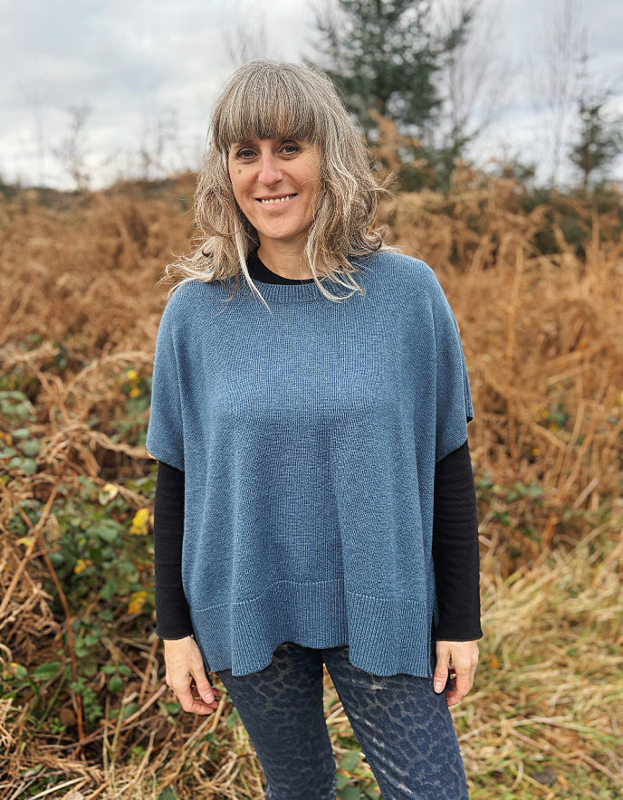 Woman wearing a blue poncho standing in a field with dry grass and trees in the background