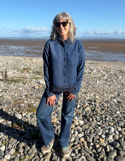 A woman with gray hair and sunglasses stands on a pebble beach wearing a dark blue denim button-up shirt and matching wide-leg jeans, with water and sky in the background.